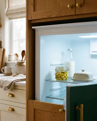 An open refrigerator with a small lidded glass jar of peeled garlic cloves in golden olive oil on a glass shelf, beside a bottle of milk and a covered butter dish.