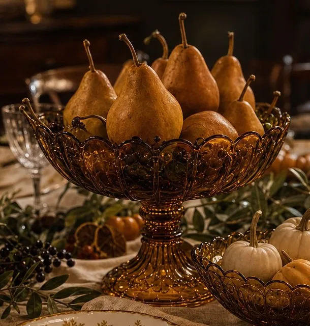 Close view of a footed amber pressed-glass compote piled with golden pears, with a lower amber bowl of small white pumpkins and olive branches beside it.