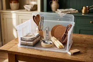 A hosting kit in a clear storage tote: cloth napkins tied with twine, wooden spoons and tongs in a jar, an instant-read thermometer, cork trivets, an extension cord, taper candles, masking tape and a marker.