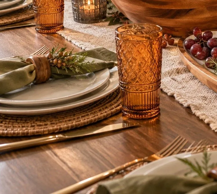 The Collected Botanical Table up close: an olive napkin in a wooden ring with a sprig of greenery on pale plates and a woven charger, an amber patterned glass, gold flatware and wooden serving pieces behind.
