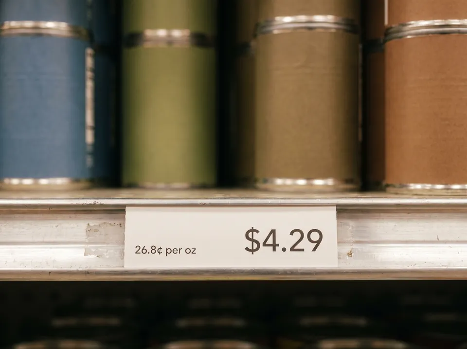 A white grocery shelf tag showing a price of $4.29 on the right and a unit price of 26.8 cents per ounce on the left, below cans with plain colored labels.