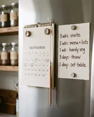 A paper calendar on a refrigerator door beside a card reading 3 wks invite, 2 wks menu and lists, 1 wk hardy veg, 3 days thaw, 1 day set table.