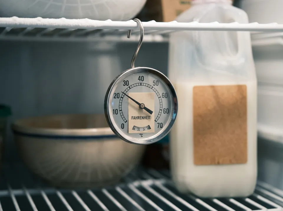 A small dial appliance thermometer hanging from a wire shelf inside a refrigerator, a carton of milk softly out of focus behind it.
