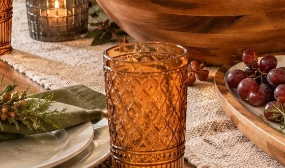 An amber patterned glass in front of a smoke-gray glass votive with a lit candle, a broad wooden serving bowl and a wood platter of red grapes on a fringed woven runner.
