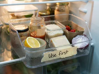 A clear refrigerator bin labeled Use first holding half a lemon, a jar of salsa, leftover rice, a wedge of cheese, strawberries and half an onion.