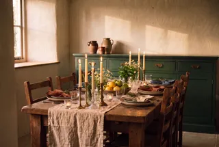 A farmhouse table built in layers with stacked cream and sage plates, mismatched napkins tied with rosemary, a low centerpiece of fruit and herbs, and lit candles at different heights.