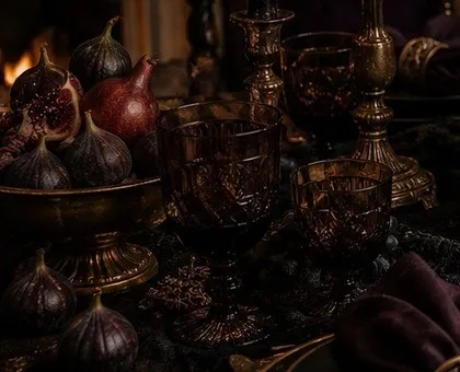 Low clusters of whole and halved figs, red pears and a brass footed bowl on a dark table beside faceted smoke-glass goblets.