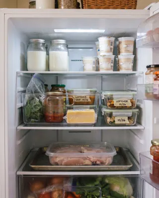 An open refrigerator with clear bins of leftovers labeled with dates, jars of milk and yogurt cups on the top shelf, and chicken on a tray below.